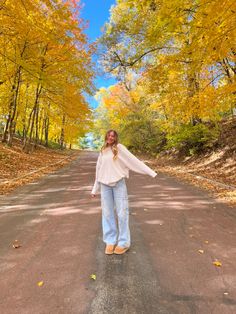 a woman standing in the middle of an empty road surrounded by trees with yellow leaves