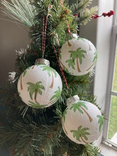 three ornaments hanging from a christmas tree in front of a window with pine needles and palm trees