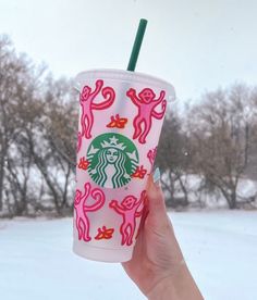 a hand holding up a starbucks cup with pink and green designs on it, in front of snow covered trees