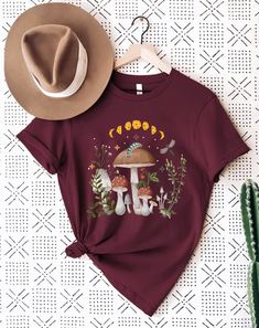 a maroon t - shirt with mushrooms and plants on it, next to a hat