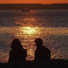 two people are sitting on the shore watching the sun go down