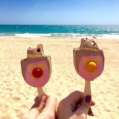 a hand holding two popsicle shaped like animals on the beach with an ocean in the background