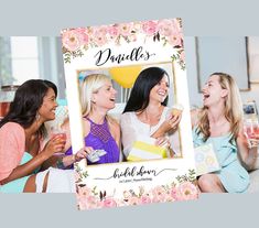 two women laughing and holding drinks in front of a photo frame with flowers on it