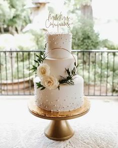 a three tiered wedding cake with white flowers and greenery sits on a gold pedestal