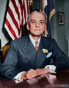 a man in a suit sitting at a table with an american flag behind him