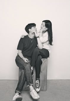 a man and woman sitting on top of a wooden bench next to each other in front of a white wall