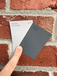 a hand is holding some paint samples in front of a brick wall that has been painted gray