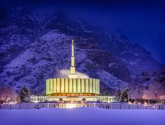 a large building with a tall steeple lit up in the snow at night, surrounded by snowy mountains