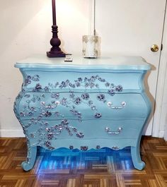 a blue dresser with flowers painted on the top and bottom, next to a lamp