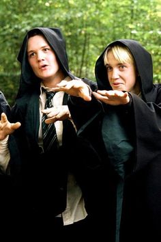 two women dressed in robes and hoods are pointing their hands at the camera with trees in the background