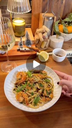 a person holding a plate of pasta with shrimp and lemons on the table next to some wine glasses