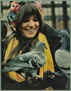 a woman with a bird on her head smiles at the camera while standing next to pigeons