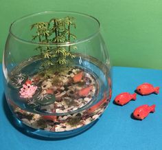 a fish bowl filled with rocks and plants next to small red plastic toy animals on a blue surface