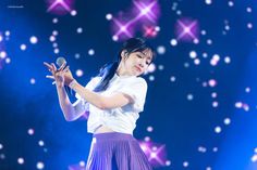 a woman in a white shirt and purple skirt holding a microphone on top of a stage