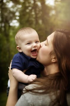 a woman holding a baby in her arms and kissing it's face with trees in the background