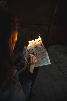 a woman sitting in front of a fire holding a piece of paper with writing on it