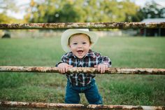 a little boy wearing a cowboy hat standing in front of an old metal fence with his hands on the bars