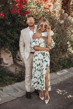 a man and woman standing next to each other in front of some bushes with red flowers