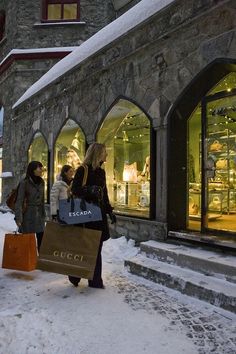 two women carrying shopping bags walking down the snow covered sidewalk in front of a store