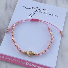 a pink bracelet with gold beads and a heart charm on the end is sitting next to a card