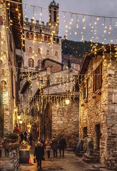 people are walking down an alley way with lights strung from the buildings