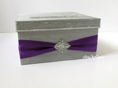 a gray box with a purple ribbon around the bottom and a diamond brooch on top