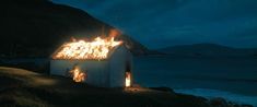 a house on fire in the dark by the ocean with mountains and water behind it