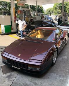 a man standing on the hood of a maroon sports car at a gas station next to other cars