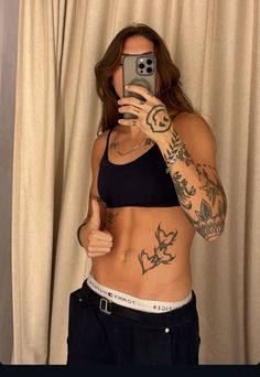 a woman with tattoos taking a selfie in front of a curtain holding a cell phone