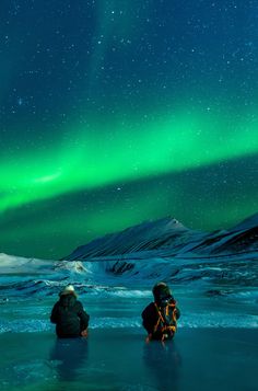 two people standing in the snow looking at an aurora bore over mountains and ice floes