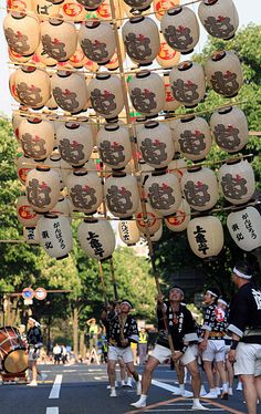 a group of people walking down a street next to a tall pole with lots of lanterns hanging from it's sides