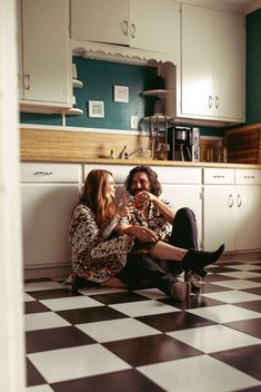 young, newly engaged couple are sitting side-by-side in their kitchen on a black & white checkered tile. They're smiling and toasting their drinks together.