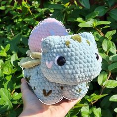 a hand holding up a crocheted stuffed animal in front of some bushes and leaves