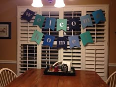 a welcome home sign hanging from the ceiling above a wooden table with chairs in front of it