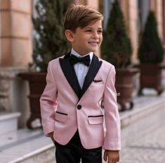 a young boy in a pink suit and bow tie standing on the sidewalk with his hands in his pockets