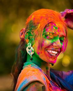 a woman with bright paint on her face and hands, smiling at the camera while she is covered in colored powder