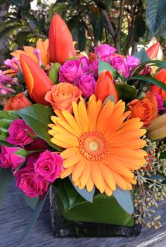 an orange and pink flower arrangement in a square vase on a wooden table with greenery