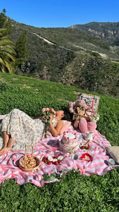 two women sitting on a blanket in the grass eating cake and drinking tea with mountains in the background