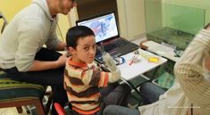 a young boy sitting in front of a laptop computer while being examined by an adult