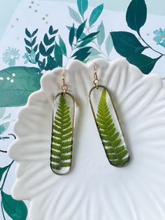 a pair of fern leaf earrings on a white plate with green leaves in the background