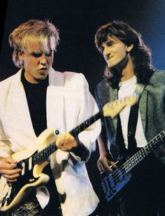 two young men are playing electric guitars on stage, one is wearing a white jacket