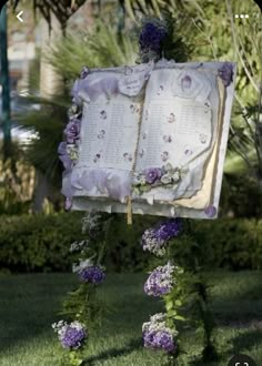 an open book decorated with purple flowers and greenery in front of some bushes on the grass