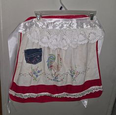 a white and red apron hanging on a door with an embroidered rooster in the pocket