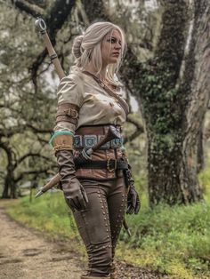 a woman dressed in costume walking down a dirt road with trees behind her and holding a stick