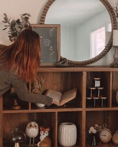 a woman reading a book in front of a mirror and shelves with figurines