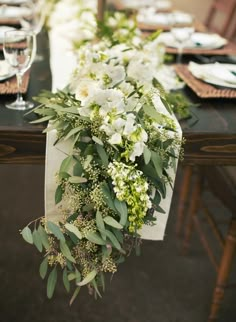 a long table with white flowers and greenery on it, along with place settings