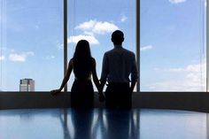 a man and woman standing in front of large windows looking out at the city skyline