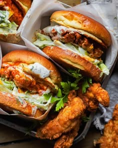 four chicken sandwiches in take out containers on top of a wooden table next to fried food