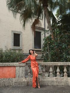 a woman in an orange dress leaning against a stone wall with palm trees behind her