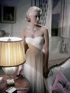 a woman in a white dress standing next to a table with a lamp on it